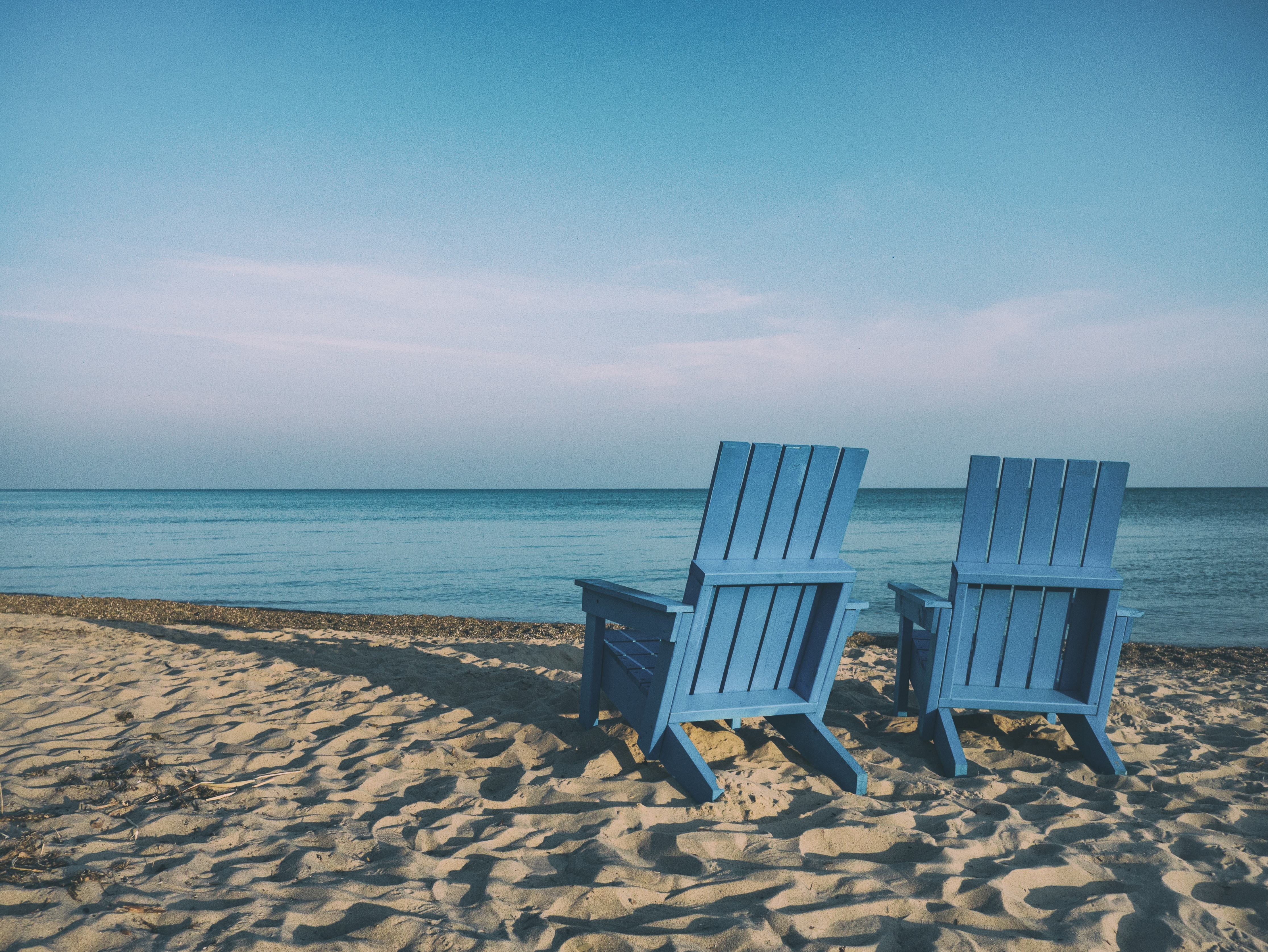 Two blue colored wooden chairs on the beach facing the clear water and light colored sky